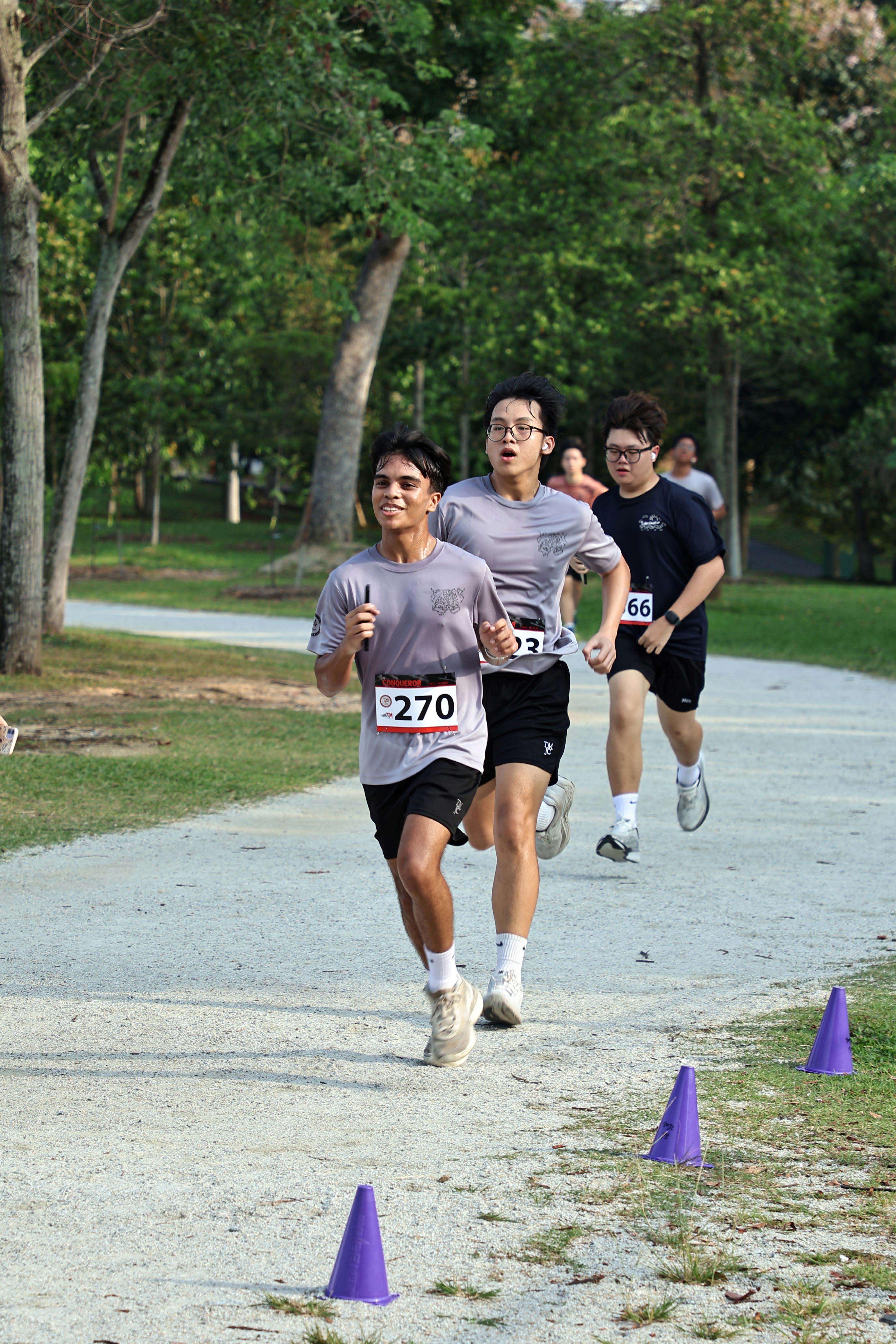 Three male students in grey T-shirts and race bibs run along a path lined with purple cones, with the lead runner smiling as he nears the finish.